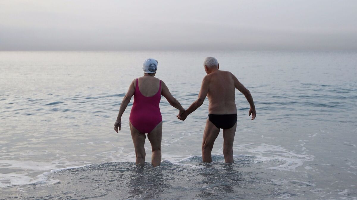 View from behind of two senior citizens, a woman in a swim cap and pink swimsuit and a man in black swim briefs, stand in the ocean, holding hands.