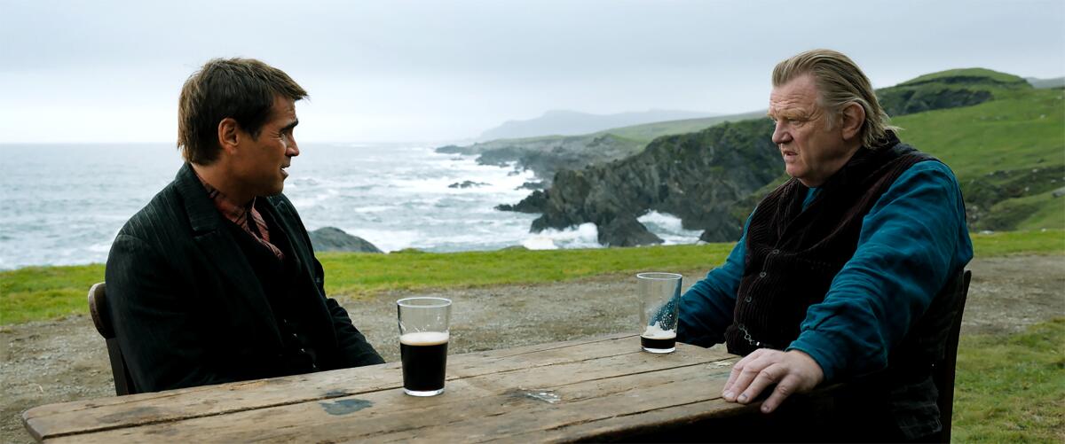 Colin Farrell and Brendan Gleeson staring at each other across a picnic table with beer glasses on it overlooking the sea