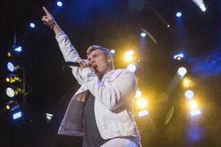 Nick Carter is wearing a white jacket and pants while performing on stage with microphone to mouth, finger pointing up