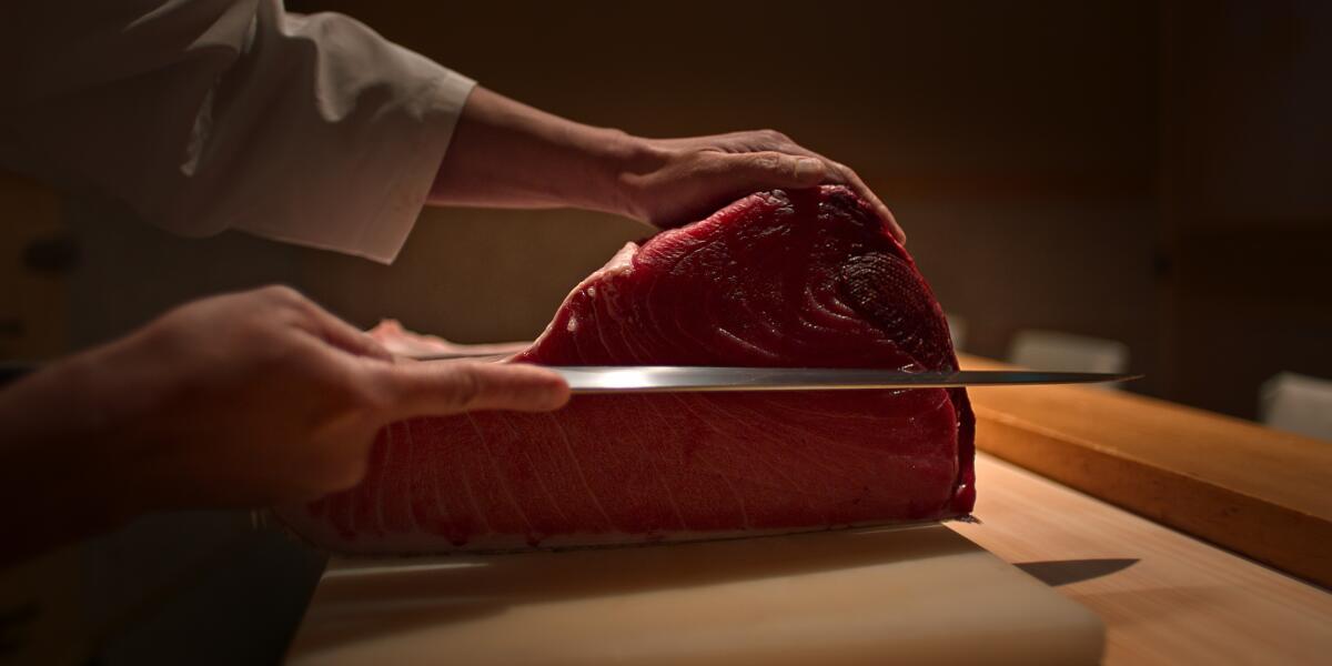 Sushi chef Takashi Saito guides a knife toward a large piece of bluefin tuna on a cutting board.
