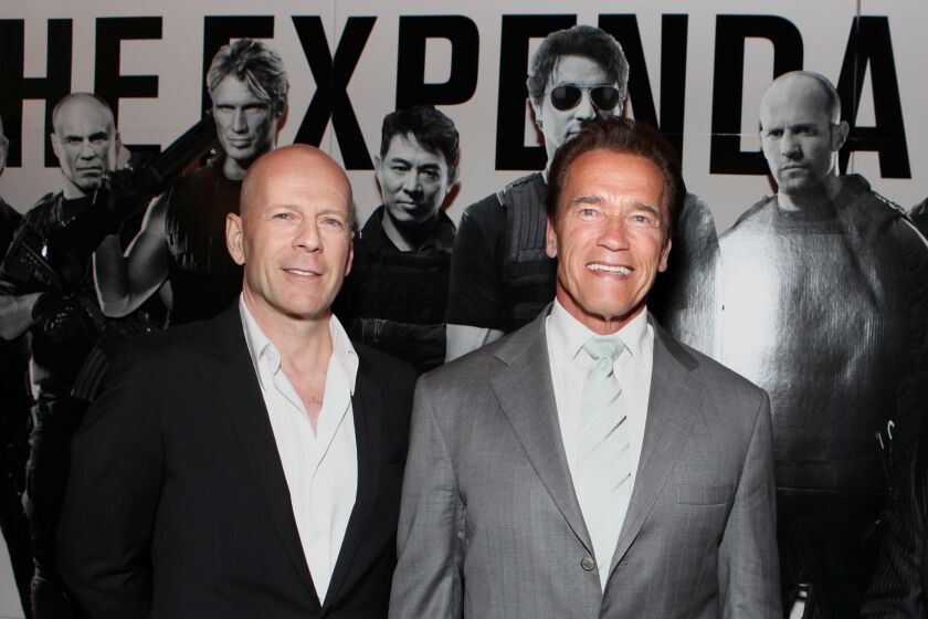 Bruce Willis in a suit shirt and black blazer standing next to Arnold Schwarzenegger in a gray suit. Both men are smiling