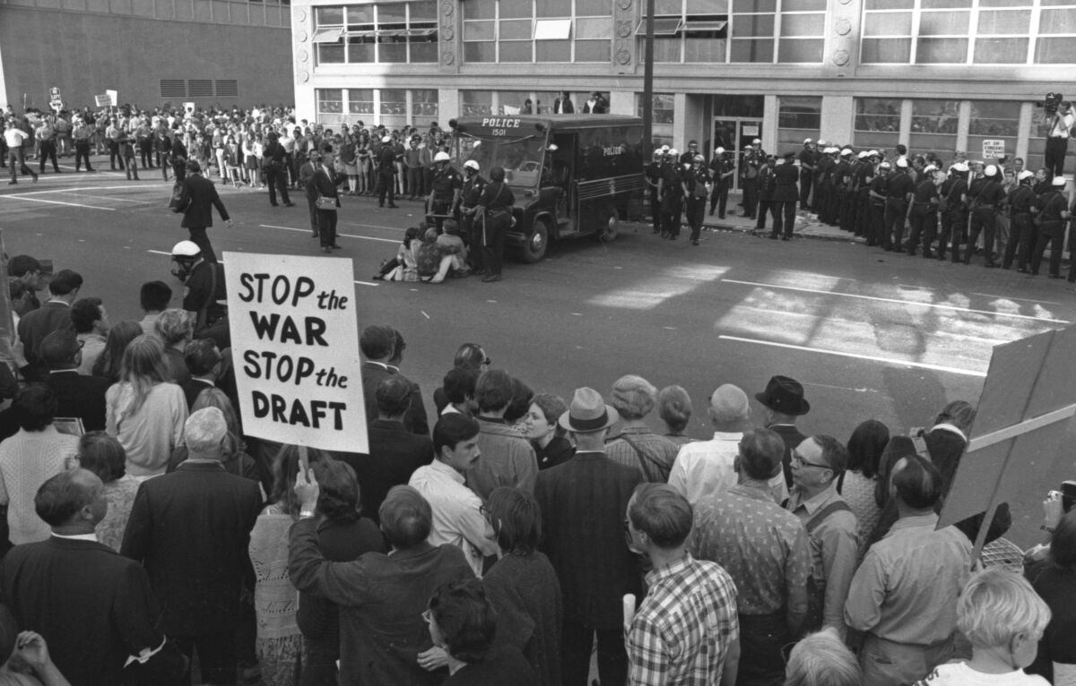 A man holds a sign that says "Stop the War. Stop the Draft" amid a crowd. Several people sit in front of a police van. 