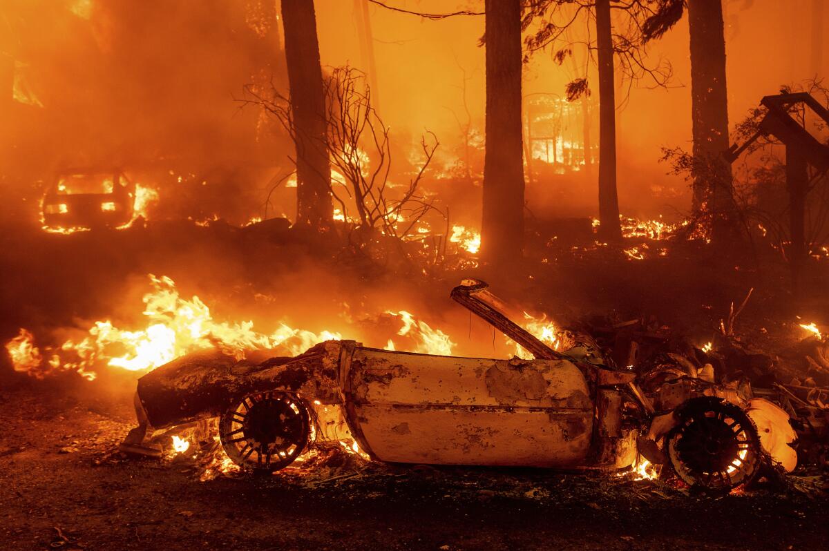 A vehicle burns in front of trees and structures on fire at night