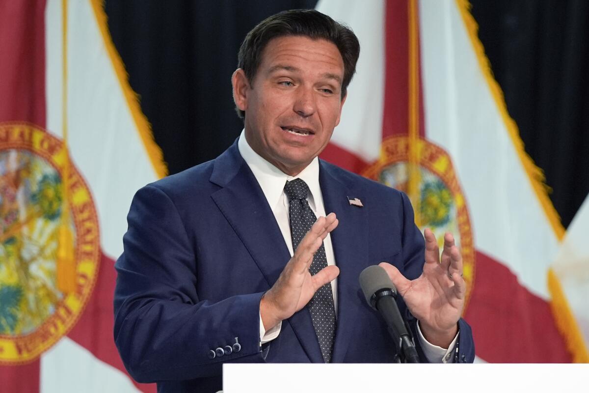 A man with dark hair, in dark suit and tie, gestures with his hands as he speaks at a news conference.