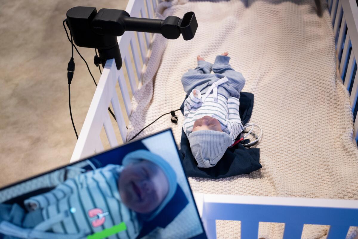 A doll lying below a baby monitor that's attached to the side of its white crib