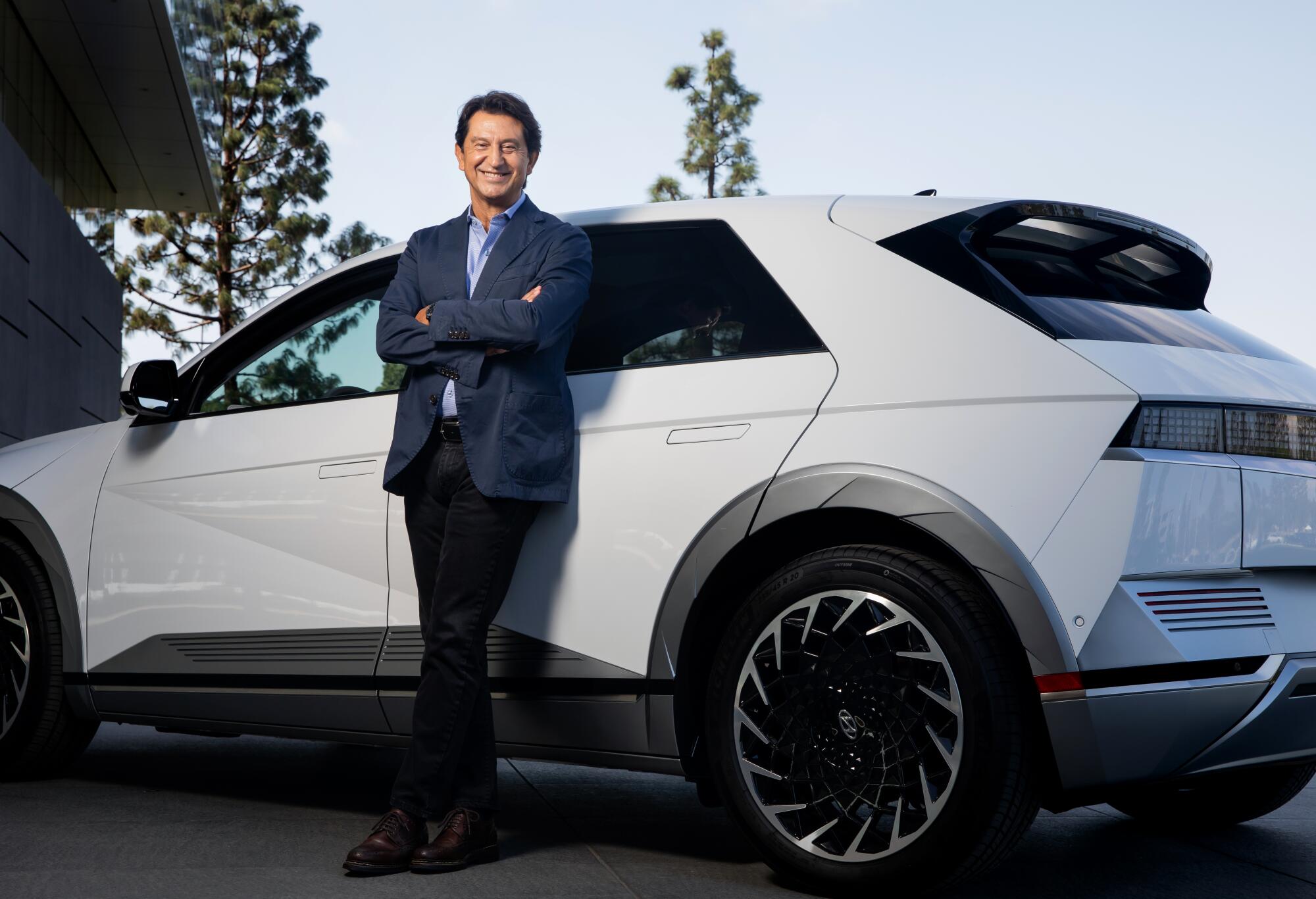 A man in a blue blazer leans against a white electric car with his arms folded across his chest.