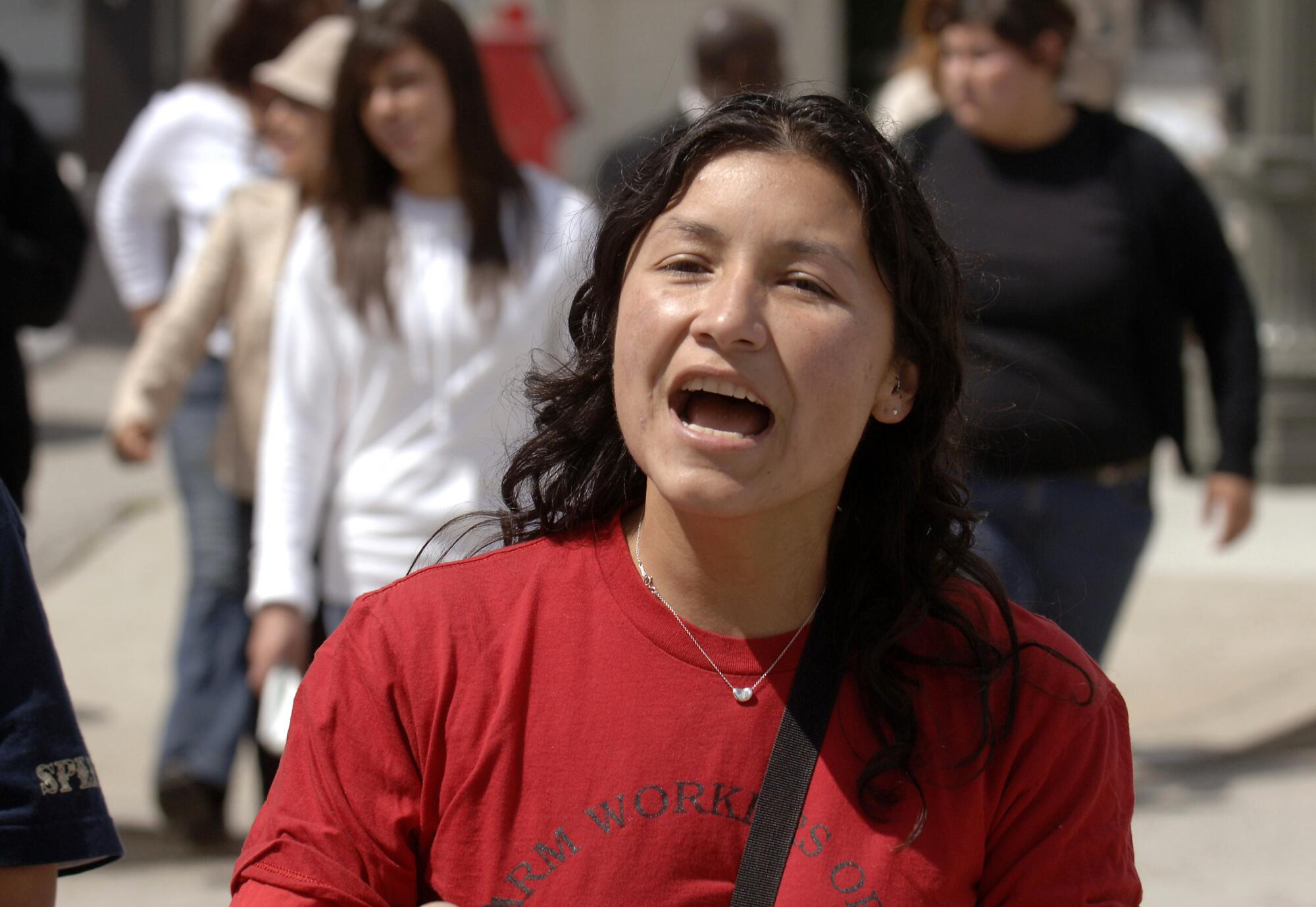 A woman with dark hair, wearing a red top, speaks as other people are seen walking behind her 