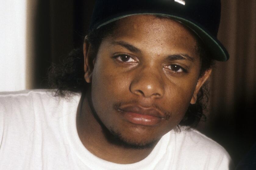 Eazy-E poses in a white t-shirt and a blue Dodgers "LA" baseball cap with a serious face