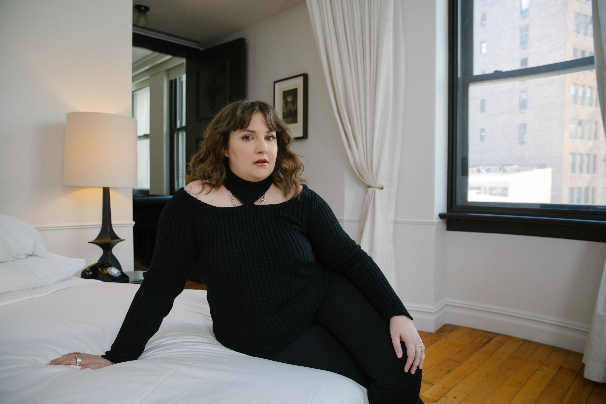 Lena Dunham sits on a white bed in a black dress for a portrait.