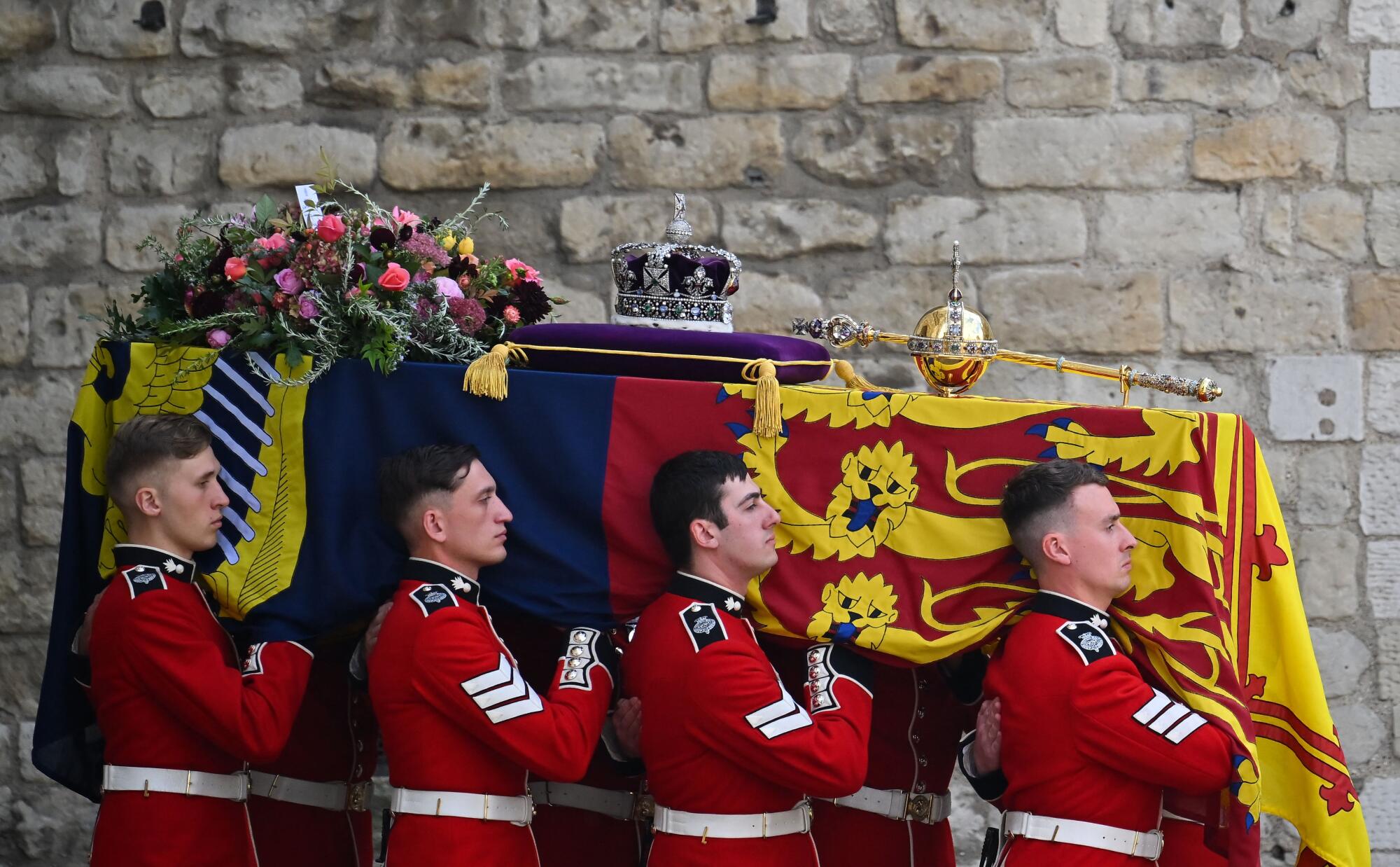 Men in red uniform carry a flag-draped coffin, which is covered with flowers, orb and sceptre and crown on a purple cushion.
