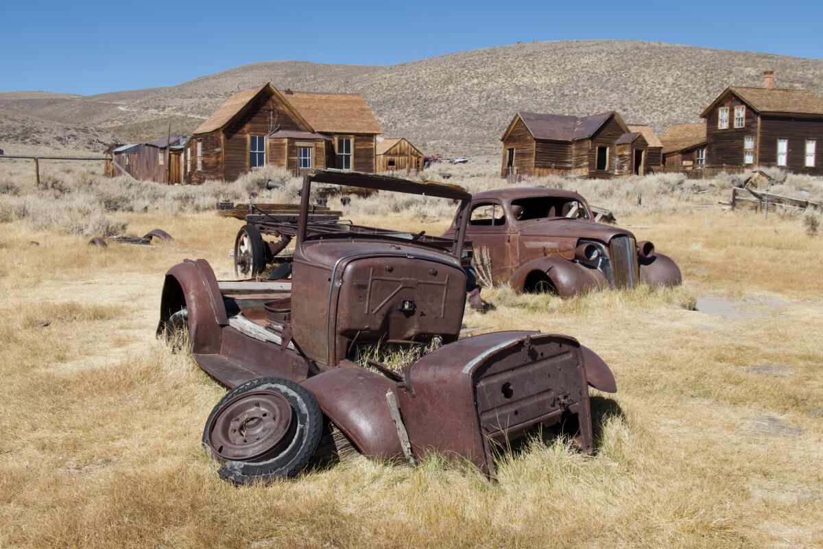 Old rusted cars lay in a dry grass and brush near old wooden houses.