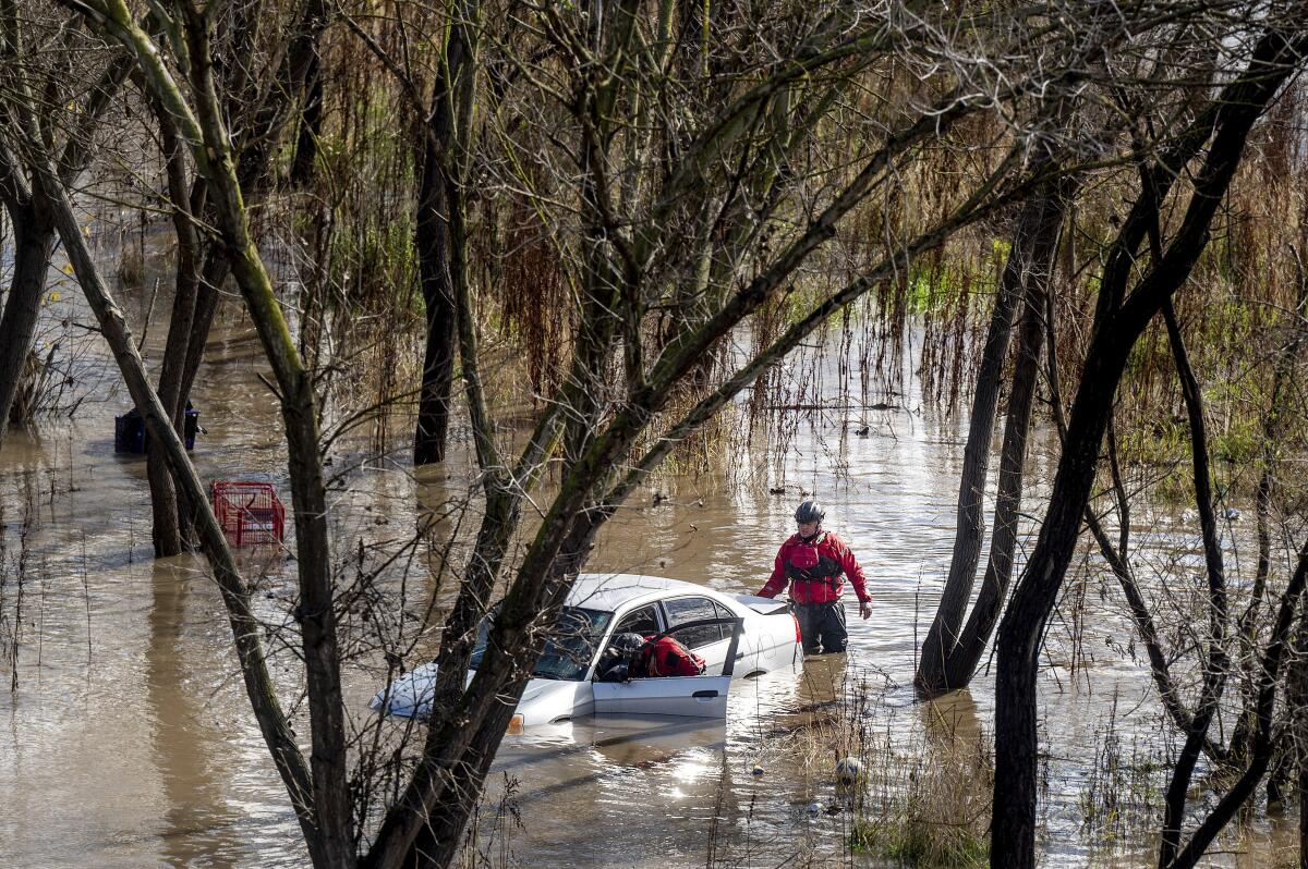 Amid trees, two people in helmets and red jackets stand in thigh-high floodwater, looking inside a submerged car.