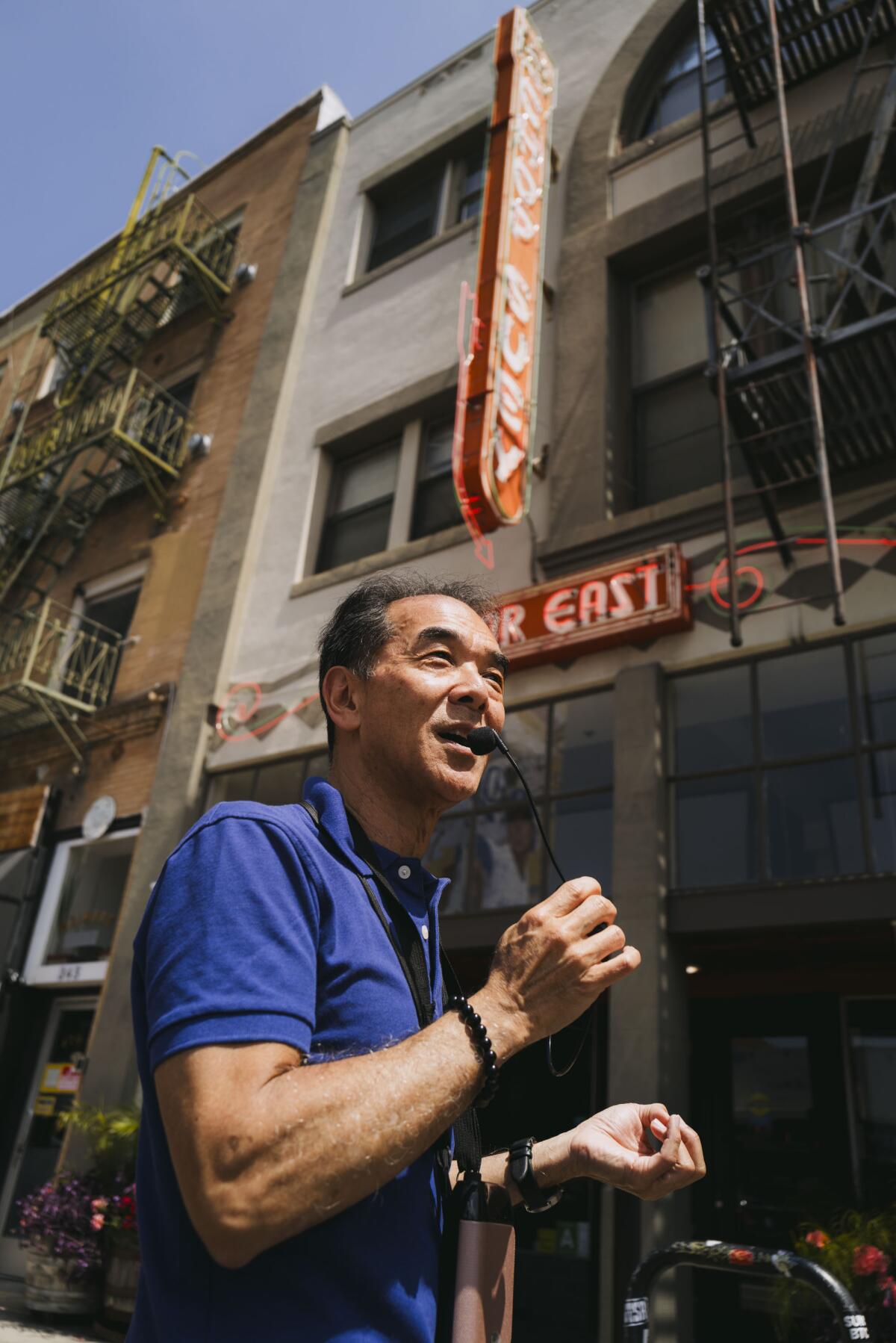 A man with a microphone walks in front of a building with a sign hanging from it.