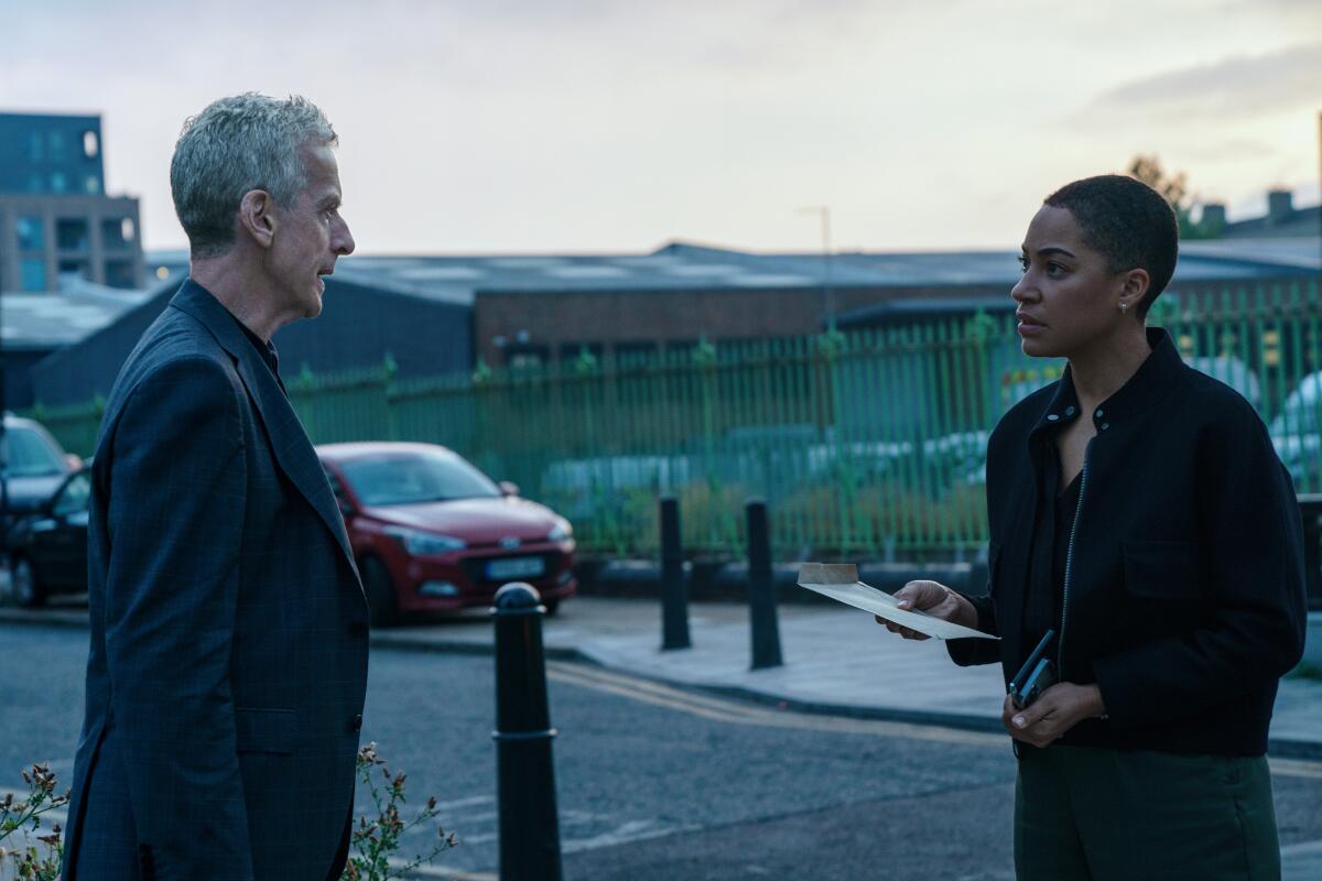 A man and a woman in dark clothing stand in a driveway across from one another.