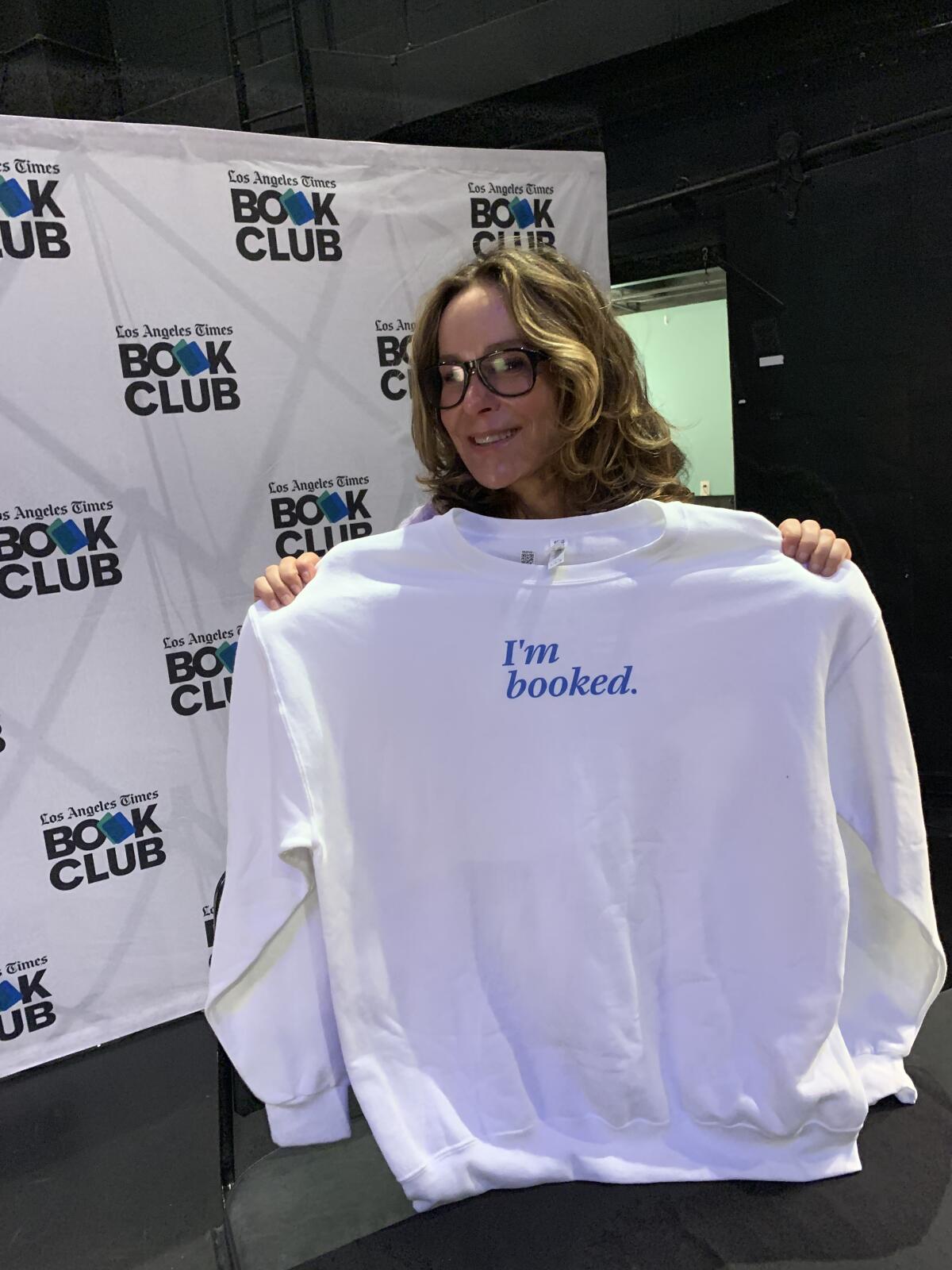 A woman wearing glasses holds up a white sweatshirt with the words "I'm Booked" on it in blue.