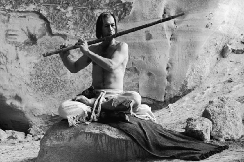 Actor David Carradine sits with his legs crossed on a blanket in a cave playing a bamboo flute.