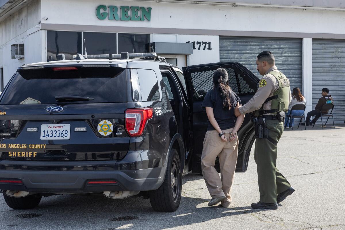 A uniformed deputy wearing a ballistic vest guides a person in handcuffs into the back of a Sheriff's Department SUV