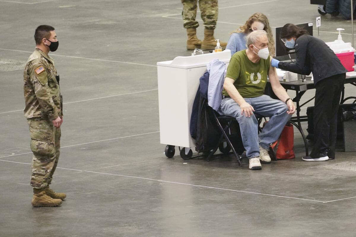 A woman gives a vaccine shot to an older man as a man in uniform stands nearby.