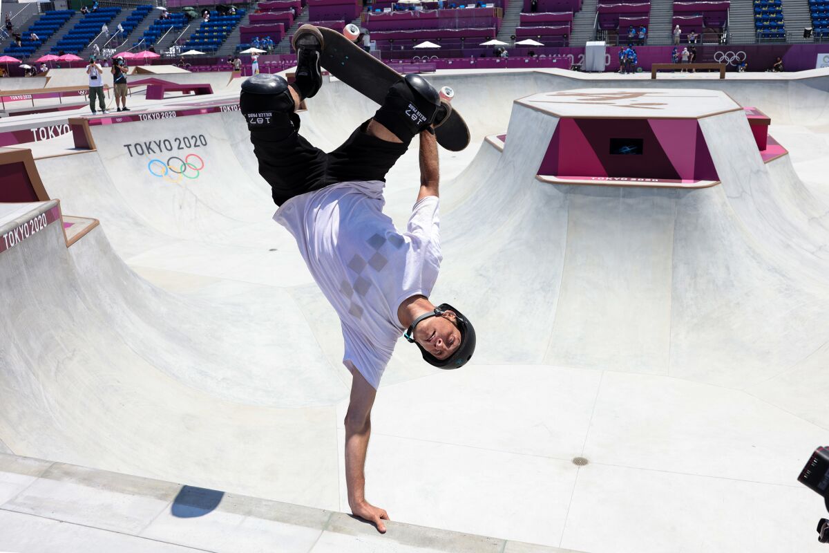 In a skating bowl, a man holds himself upside down with one arm; the other hand holds his skateboard against his feet. 