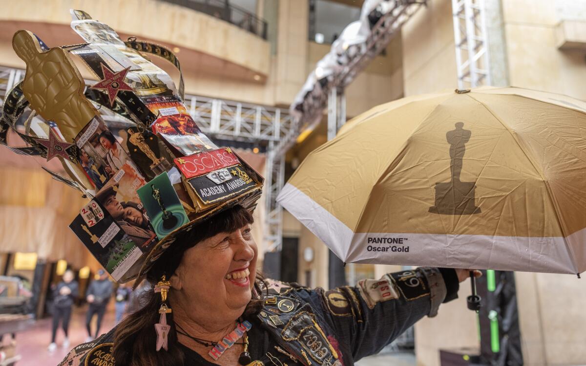 A woman in a towering hat constructed of movie scene and Oscar cutouts holds an Oscar-themed umbrella.