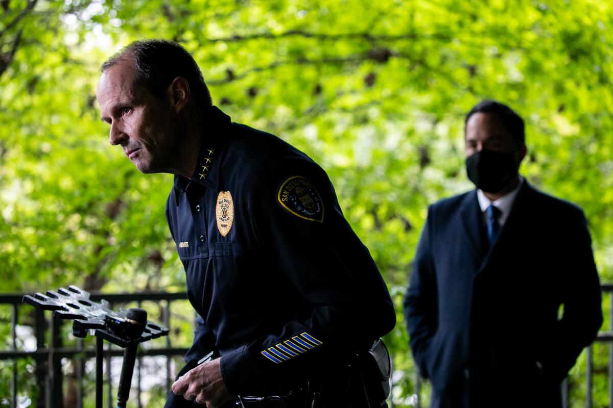 A man in a police uniform leans over a microphone as a man in a suit watches.