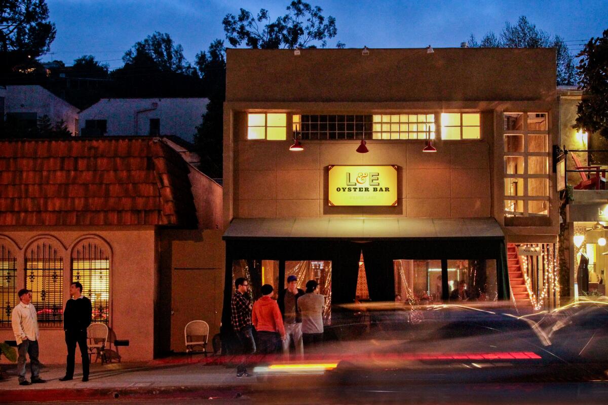 Several people on a sidewalk in front of two buildings with warm lighting after sunset; a sign on one reads "L&E Oyster Bar"