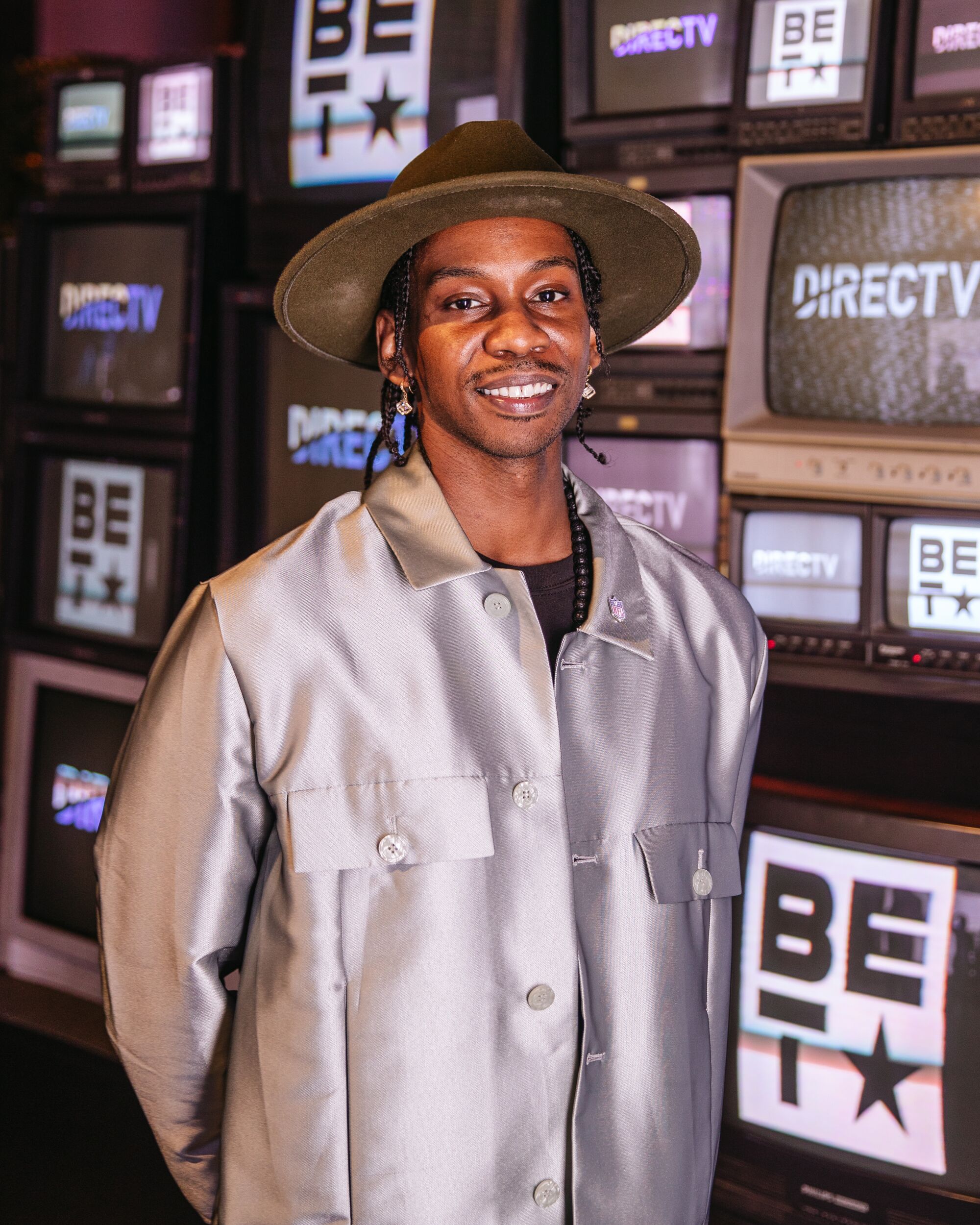A man in a loose, long-sleeve silver shirt and brown hat poses for a portrait in front of TVs.