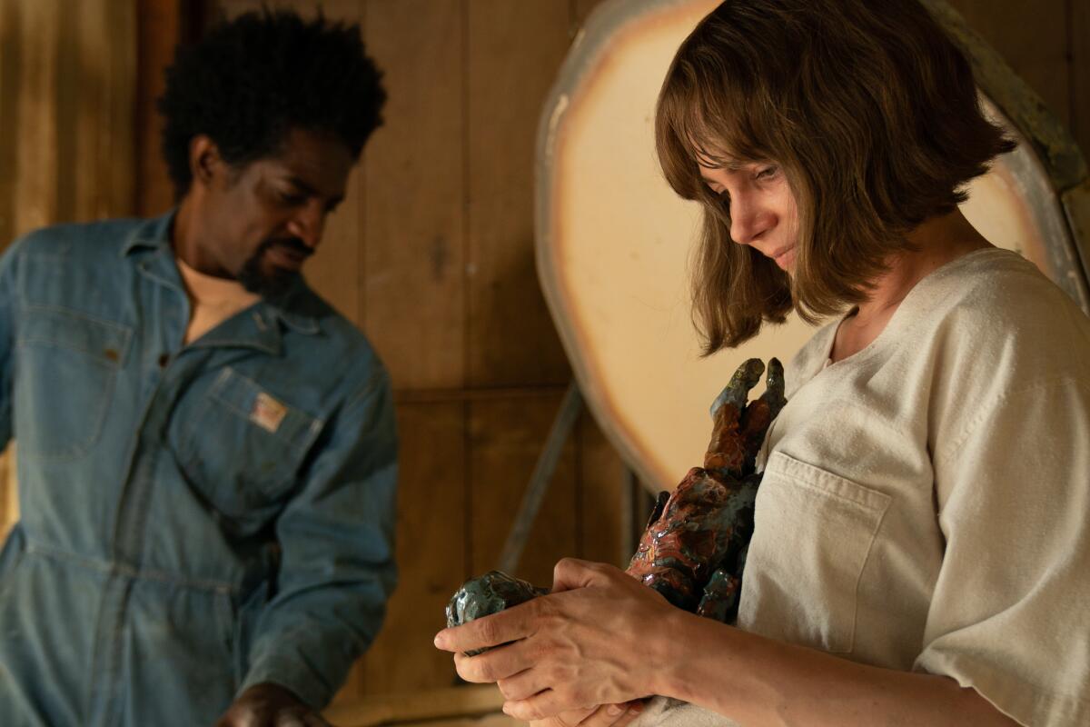 A woman works on a sculpture as a man in a blue jumpsuit stands in the background