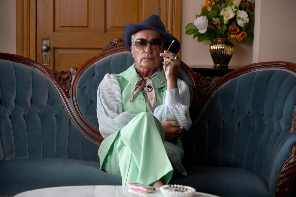 Udo Kier holds a cigarette while wearing a mint green pantsuit, sunglasses and a fedora with his legs crossed at the knee.