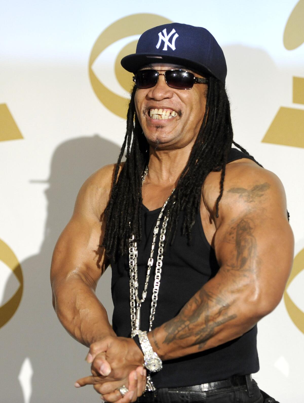 Melle Mel is posing, flexing his arms while wearing a black tank top, silver chains, a Yankees cap and dark sunglasses