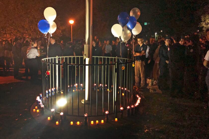 Varias personas se reúnen alrededor de un asta adornada con velas y globos de colores azul, dorado y blanco, representativos de la Escuela Secundaria Saugus, en el Parque Central de Santa Clarita, California, después de un tiroteo en la escuela, el jueves 14 de noviembre de 2019. (AP Foto/Stefanie Dazio)