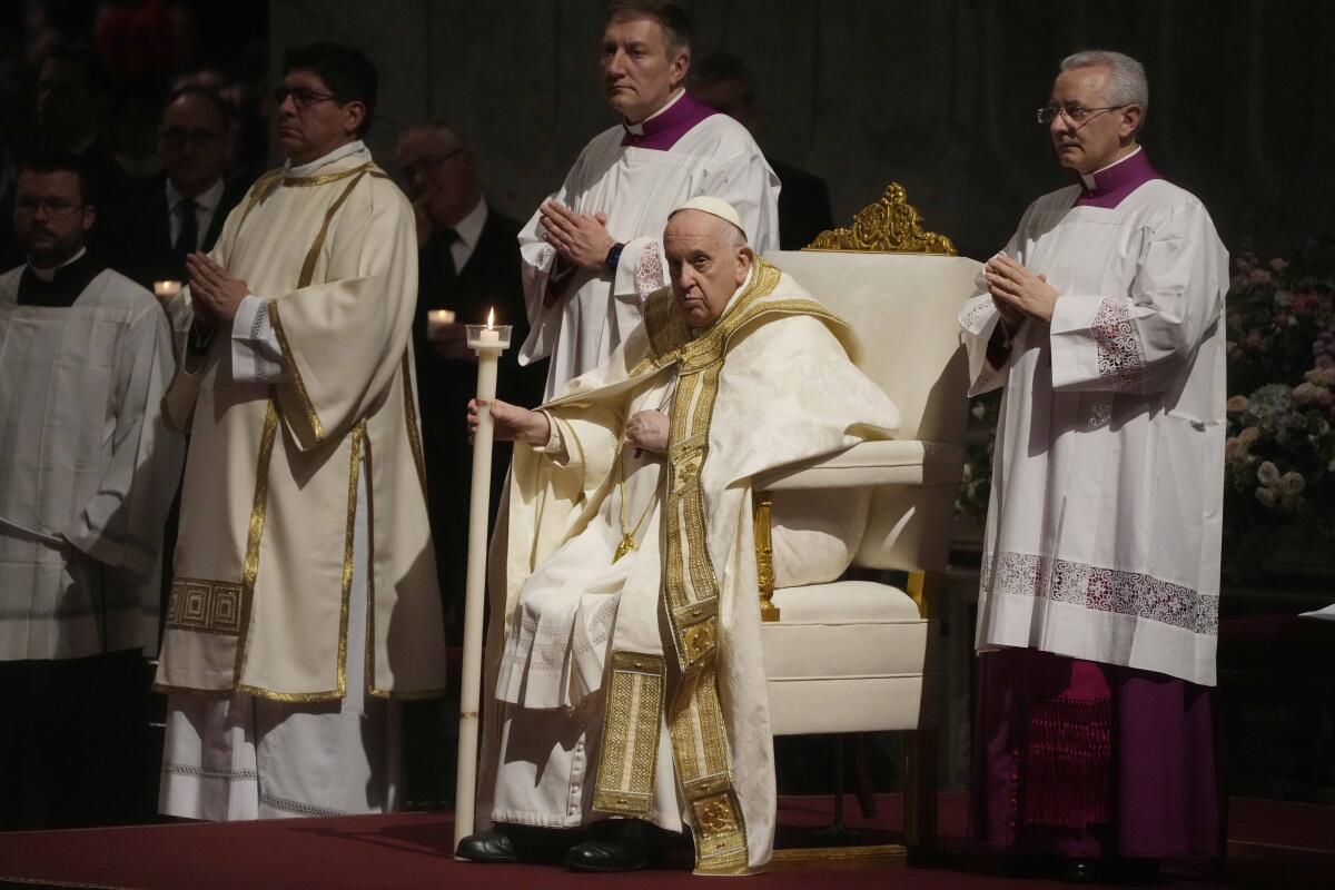Pope Francis holds a Paschal candle while sitting and flanked by men in vestments.