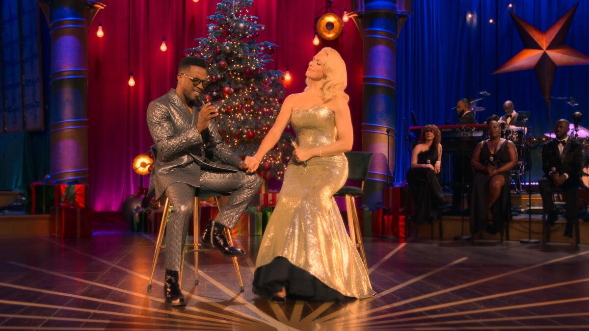 Leslie Odom Jr. and Hannah Waddingham sit on stools singing and holding hands in front of a Christmas tree.