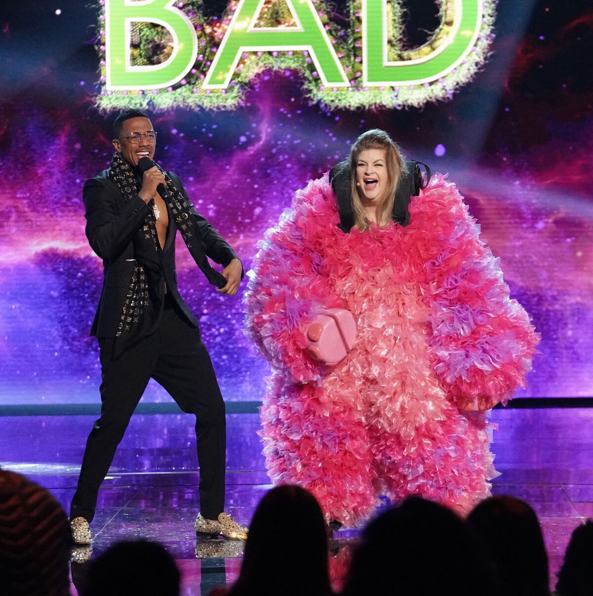 A man with a microphone stands on stage next to a woman in a fluffy pink costume