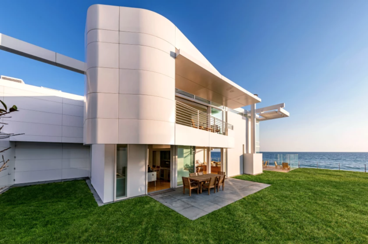 A white house with grass in front and ocean shore beyond it.