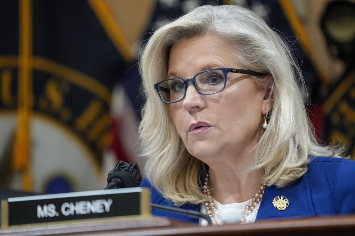 A blond woman wearing glasses sits behind a nameplate that says Ms. Cheney.