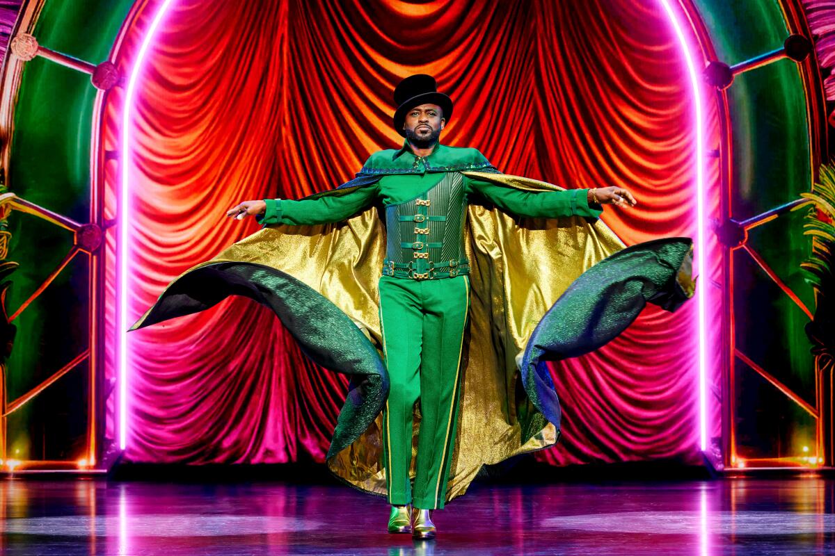 A man in a green outfit and gold-lined green cape onstage before a red draped curtain.