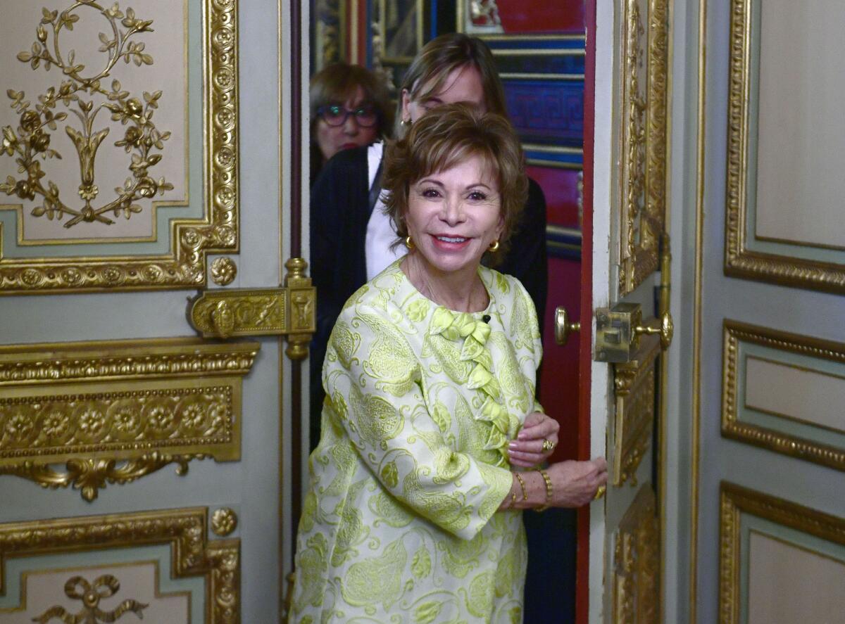 Isabel Allende, in bright floral jacket, is seen walking through a door with gilded decorative elements