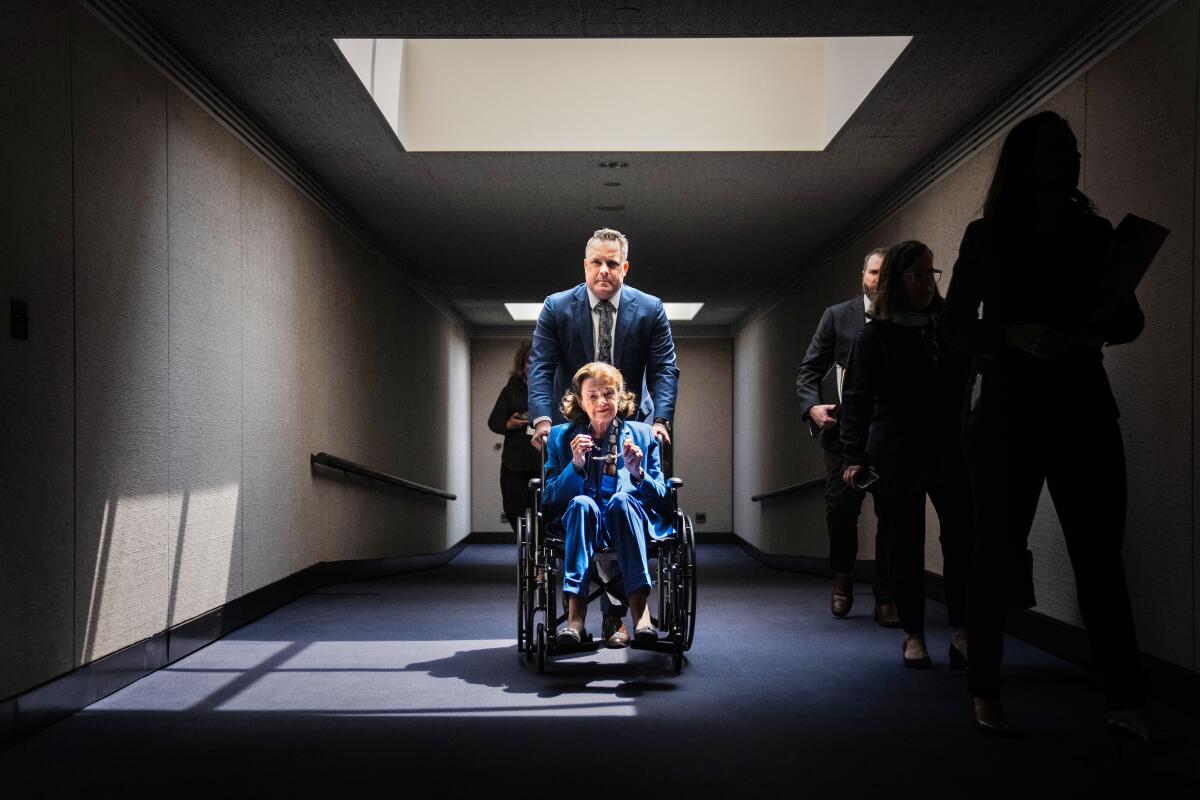 A woman is seated in a wheel chair pushed a by a man in suit and tie down a hallway lighted by a skyline. 