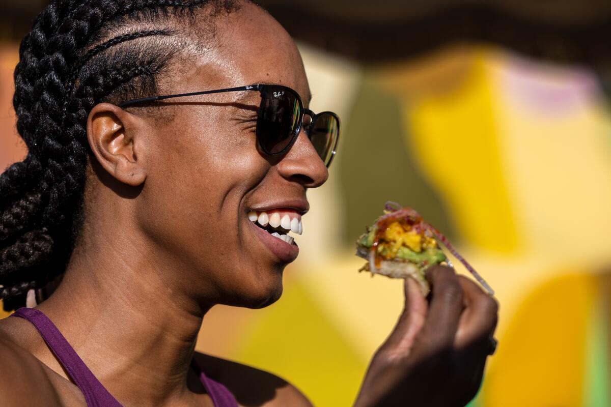 A smiling woman in sunglasses enjoys a slice of avocado toast.