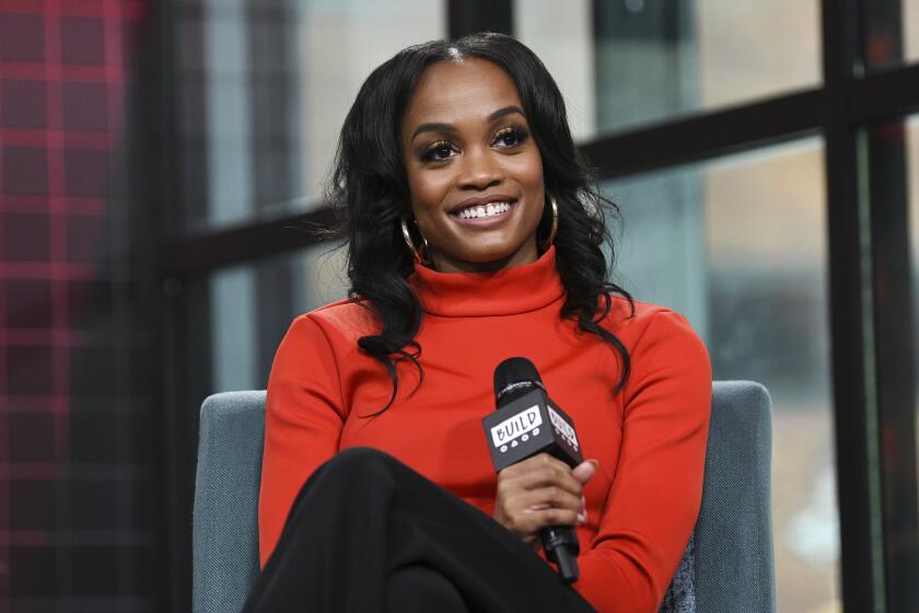 Rachel Lindsay in an orange turtleneck holding a microphone