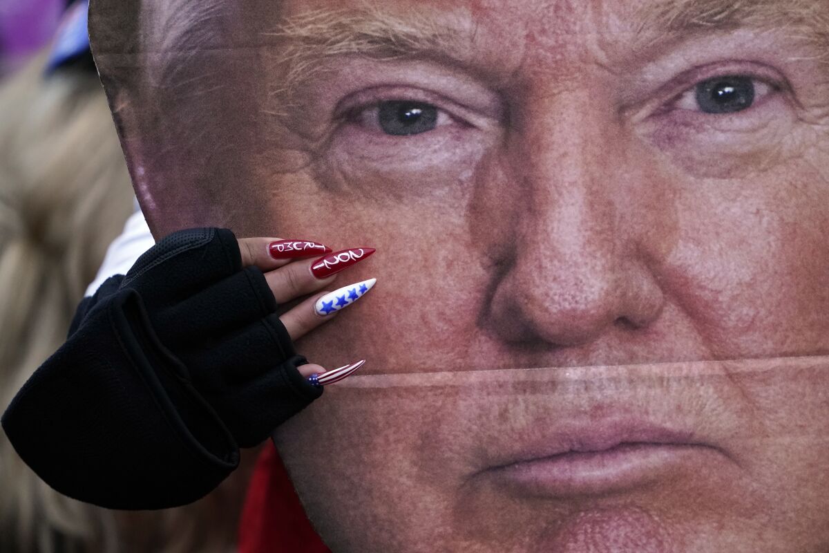 A closeup of a woman's hand, with long pointed fingernails painted with "Trump 2021," holding a cutout of Trump.