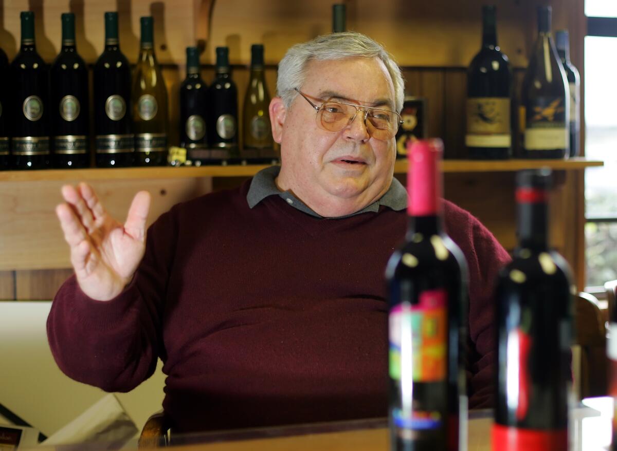 A seated man gestures in a room with many wine bottles