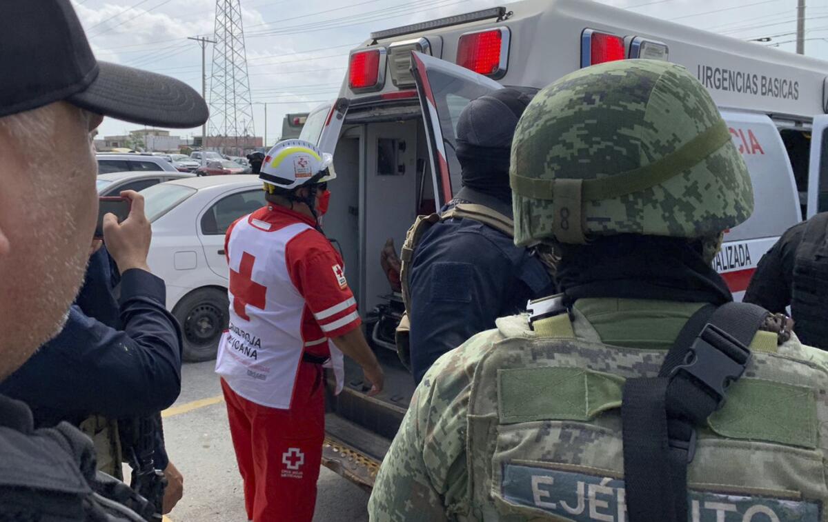 A worker in red clothing, with a white vest bearing a logo of a red cross on the back, stands facing the rear of an ambulance