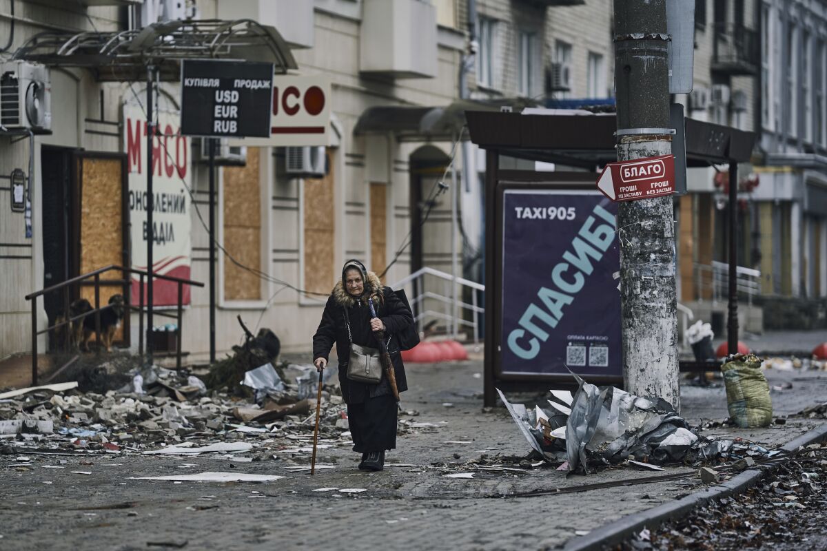 A woman walks with a cane past boarded-up windows on a sidewalk full of debris.