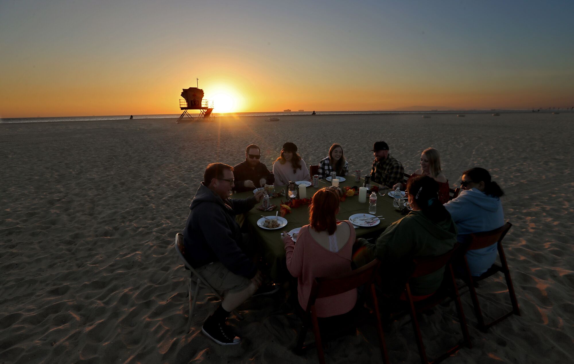 Nine people sit for dinner at a round table on the beach at sunset.