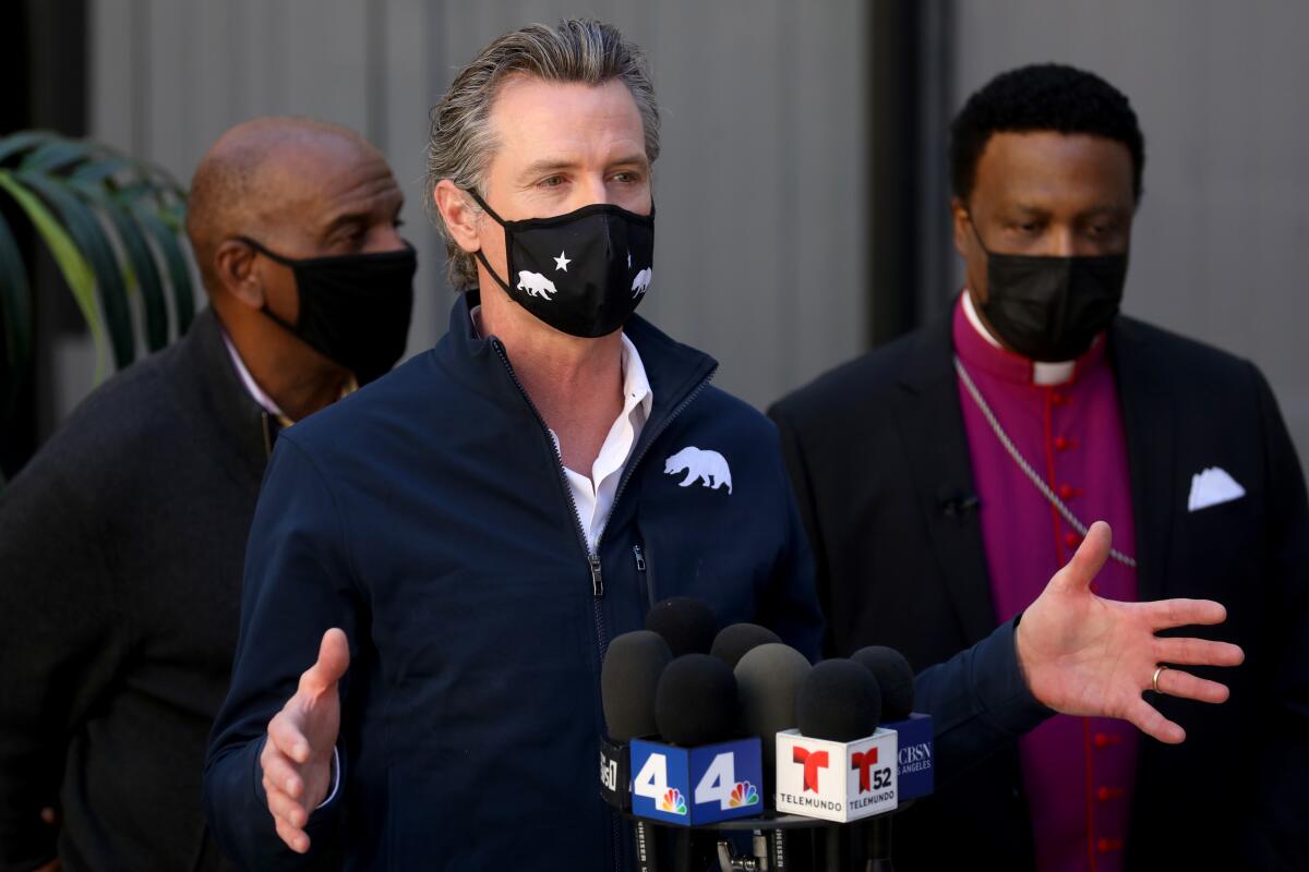 Gavin Newsom, wearing a mask, speaks into microphones with two people behind him.