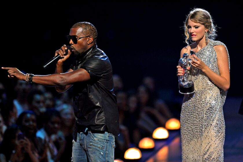 Kanye West in a black shirt, jeans and sunglasses holding a microphone on stage with Taylor Swift holding an award