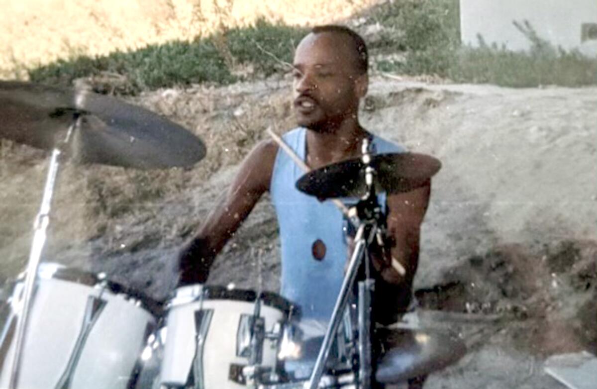 A grainy photograph of a man in a sleeveless shirt drumming at a drum set outside by the grass.