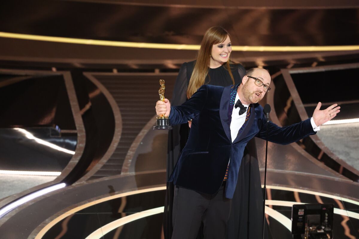 Two people stand onstage, one holding an Oscar and speaking into a microphone